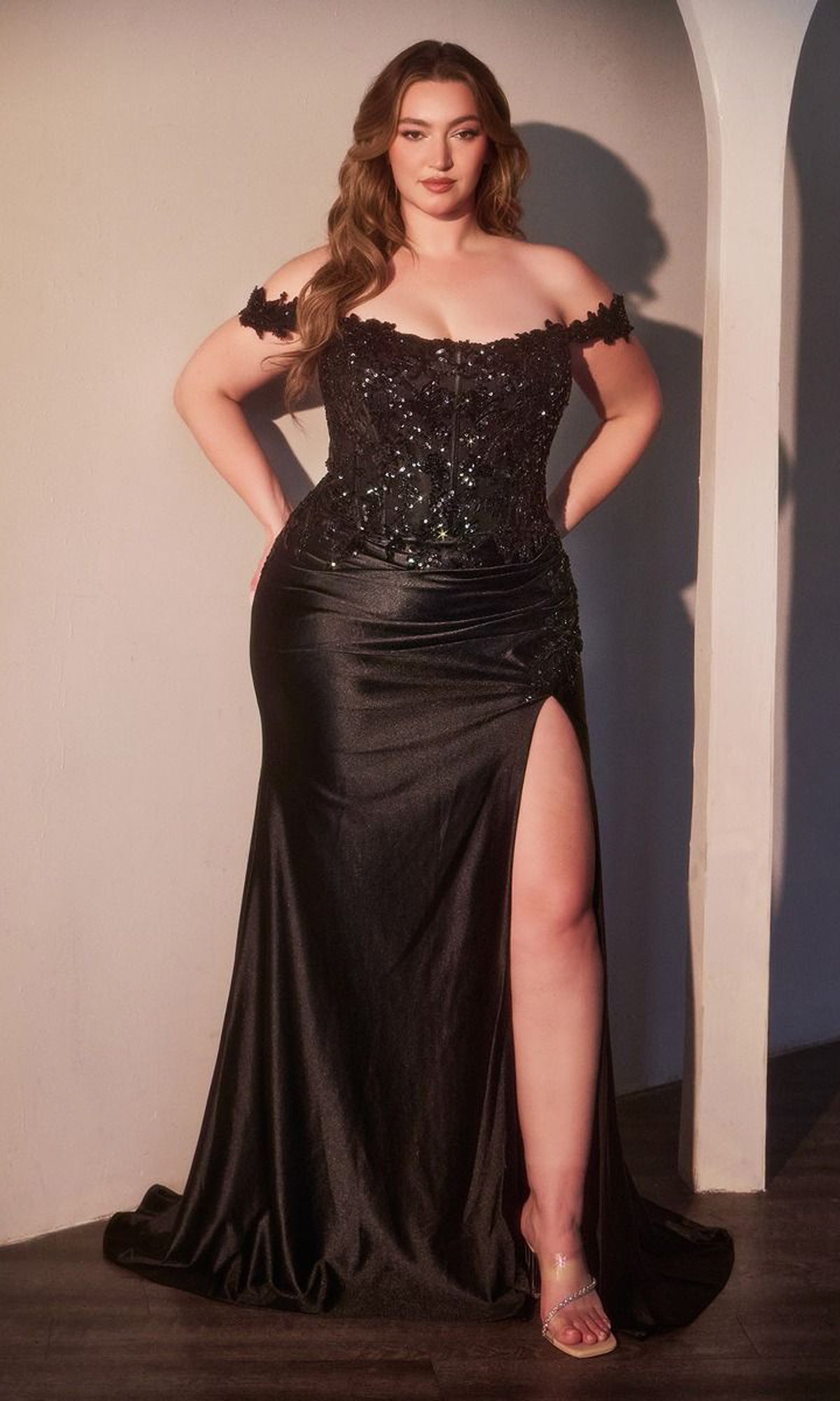 A woman poses confidently indoors with hands on her hips, wearing the Plus-Size Off-Shoulder Prom Gown: Ladivine CDS537C—an elegant black sequin evening dress with a thigh-high slit and open-toe heels.